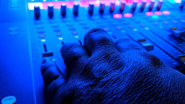 A close-up shot of a music composer in deep concentration, working at a mixing console in a dimly lit studio. The focus is on their hands and the intricate details of the equipment around them.